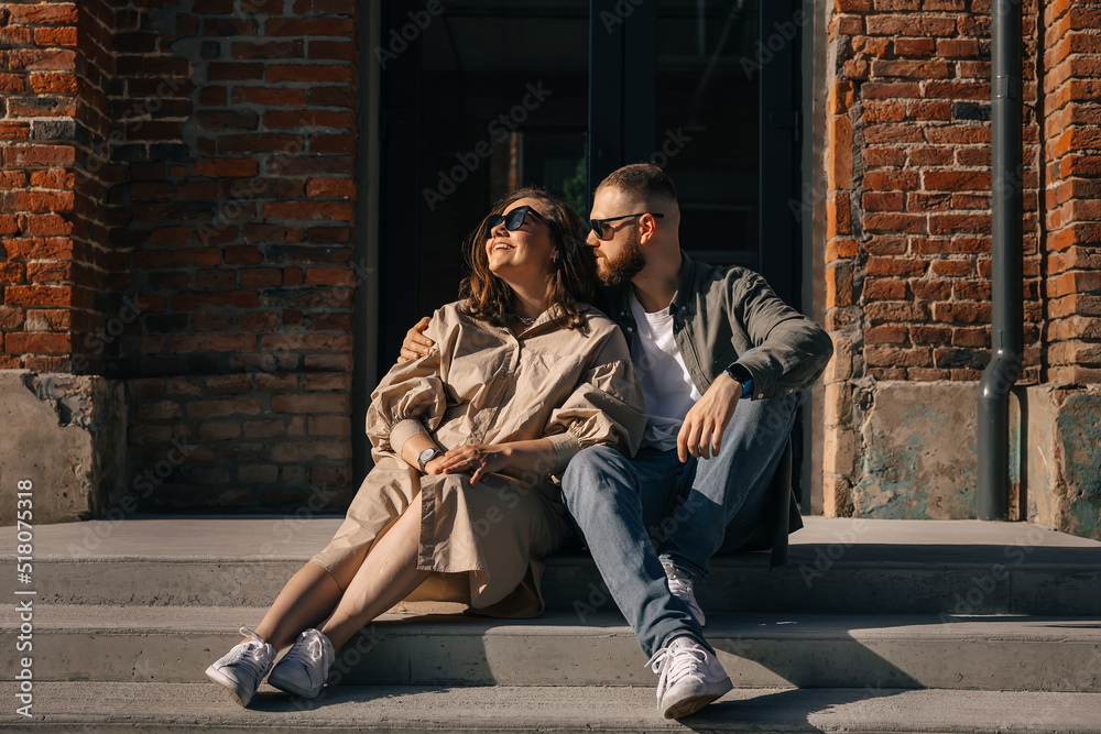 Loving and stylish couple in sunglasses sit on the stairs on the street against brick wall