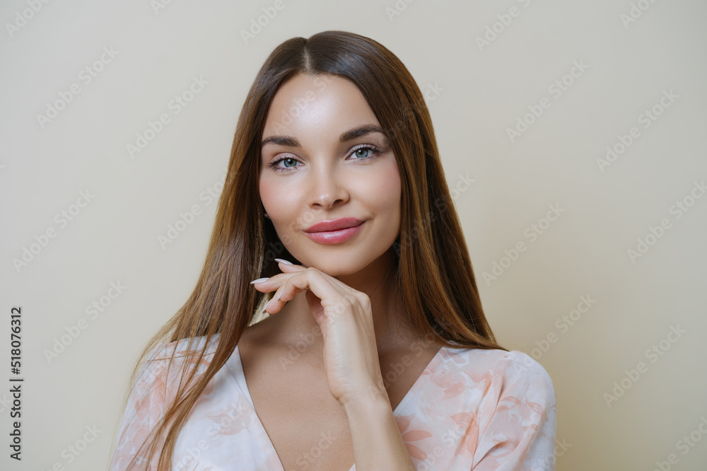 Natural beauty and people concept. Portrait of beautiful dark haired young European woman touches jawline gently, dressed in stylish dress, isolated over beige background, dreams about perfect day Natural beauty and people concept. Portrait of beautiful dark haired young European woman touches jawline gently, dressed in stylish dress, isolated over beige background, dreams about perfect day