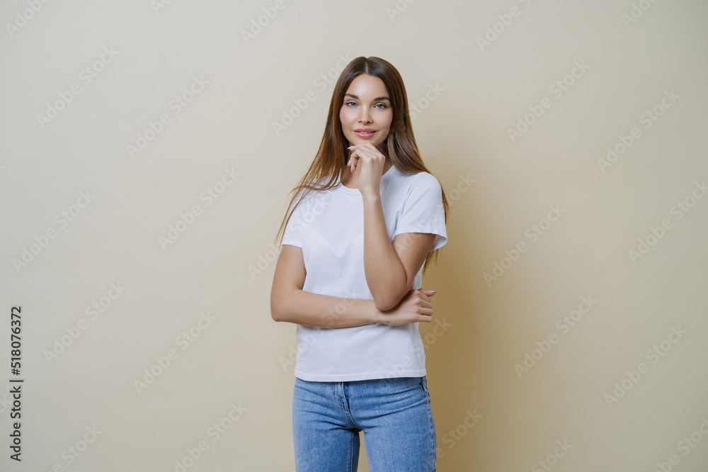 Photo of brunette young European woman holds chin, wears white t shirt and jeans, enjoys conversation with her mates, isolated over brown background. People, lifestyle, face expressions concept Photo of brunette young European woman holds chin, wears white t shirt and jeans, enjoys conversation with her mates, isolated over brown background. People, lifestyle, face expressions concept