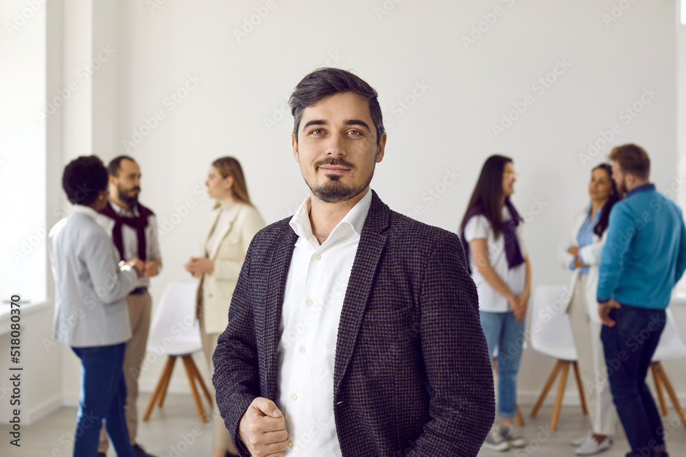 Charming businessman in a white shirt and black jacket with glasses posing in front of unfocused group of multinational people businesswoman and business man at a business meeting office space. Charming businessman in a white shirt and black jacket with glasses posing in front of unfocused group of multinational people businesswoman and business man at a business meeting office space.