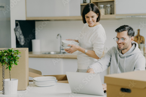 Moving house and real estate. Caring brunette woman holds pile of plates, unpacks personal belongings, husband asks her advice, chooses something to buy in internet, focused in laptop computer