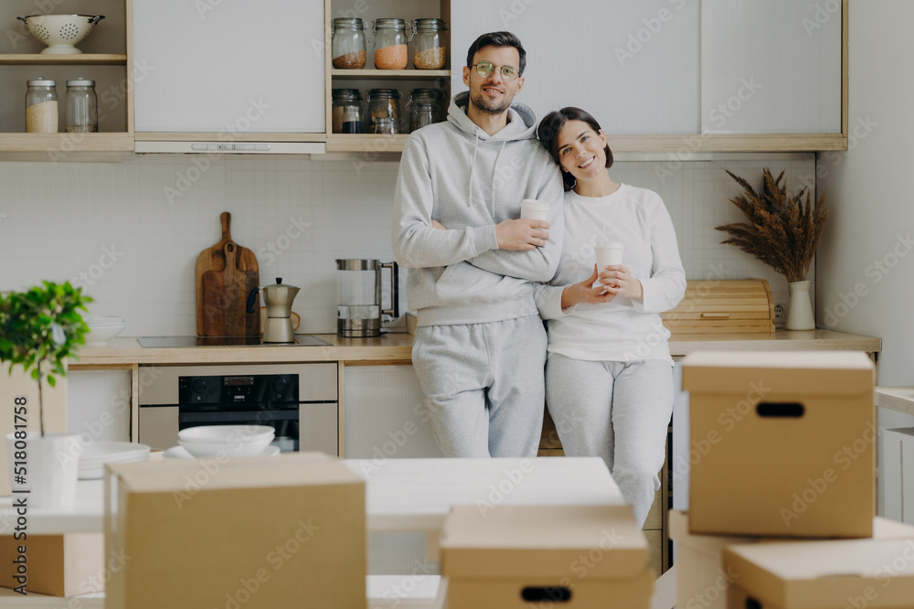 Smiling relaxed husband and wife drink coffee at kitchen, hold paper cups with hot beverage, dressed in casual outfit, relocate in new apartment, pile of cardboard boxes with property in foreground. Smiling relaxed husband and wife drink coffee at kitchen, hold paper cups with hot beverage, dressed in casual outfit, relocate in new apartment, pile of cardboard boxes with property in foreground.