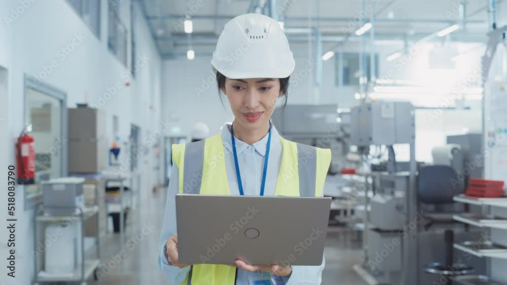 Portrait of a Happy Asian Female Engineer in Hard Hat Standing and Using Laptop Computer at Electronic Manufacturing Factory. Technician Working on Research and Development Production Plan. Portrait of a Happy Asian Female Engineer in Hard Hat Standing and Using Laptop Computer at Electronic Manufacturing Factory. Technician Working on Research and Development Production Plan.