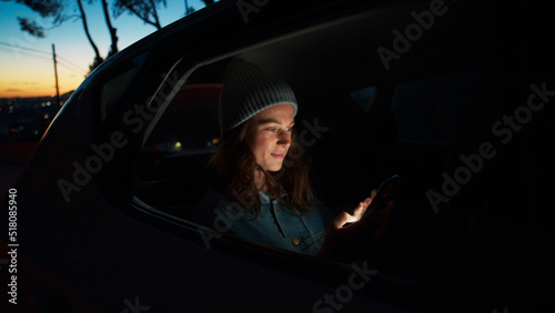 Side night cinematic view of young beautiful woman sit in taxi car back seat near window on road trip, hold, check on mobile phone, use smartphone, scroll social media news, read messages and smile