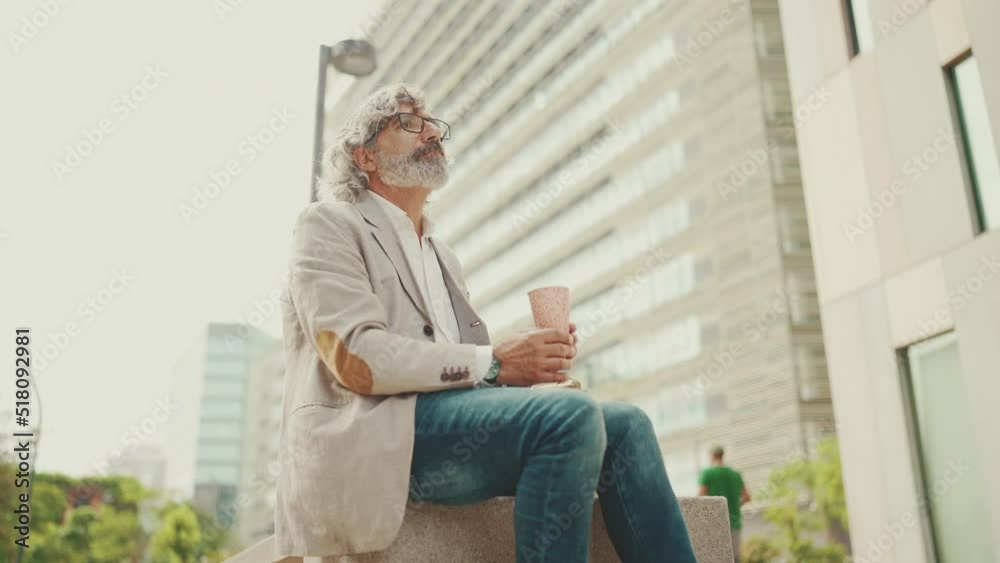 Mature businessman with beard in eyeglasses wearing gray jacket holding takeaway coffee. Middle aged manager enjoying morning coffee while sitting outside the office Mature businessman with beard in eyeglasses wearing gray jacket holding takeaway coffee. Middle aged manager enjoying morning coffee while sitting outside the office