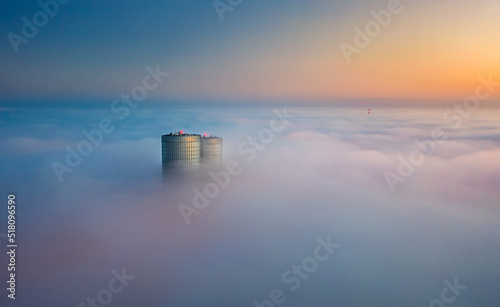Obraz na płótnie Skyscraper rooftop over the clouds at sunrise. Thick fog covers the Riga city, and warm sunlight over the clouds and church tower. w salonie