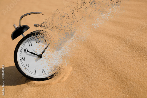 Time is running out. Black alarm clock vanishing on sand in desert