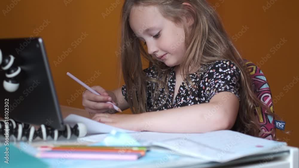 Little cute girl studying online at home using tablet. Girl writing with a pencil in her workbook. Online schooling during pandemic lockdown Little cute girl studying online at home using tablet. Girl writing with a pencil in her workbook. Online schooling during pandemic lockdown