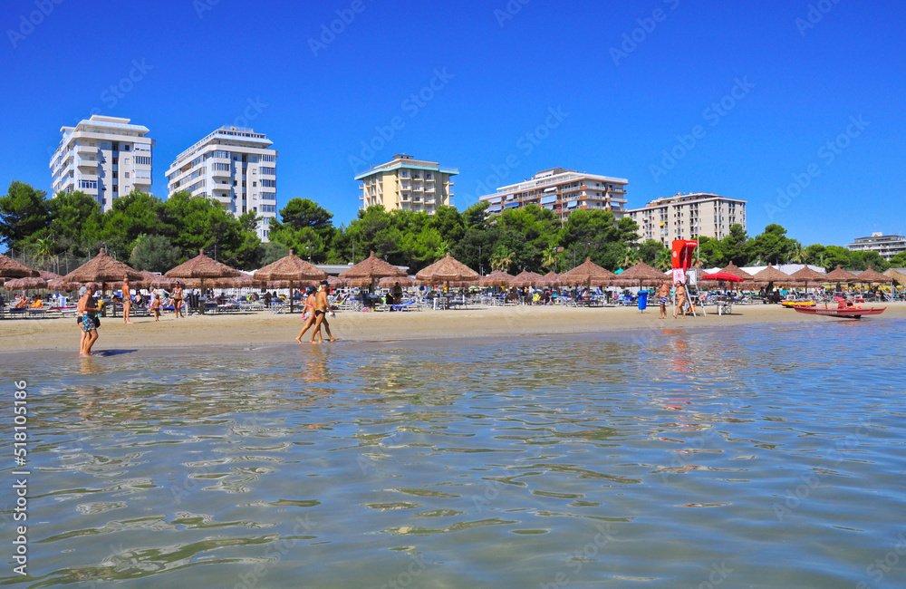 foto panoramica giornata di mare dall'acqua con vista su pineta palazzi e ombrelloni con gente che passeggia sulla riva, inizio estate