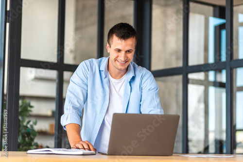 Confident entrepreneur standing in the office near the workplace and working using a laptop. Happy male young businessman with computer, looking at the screen, smiles friendly. Distant work