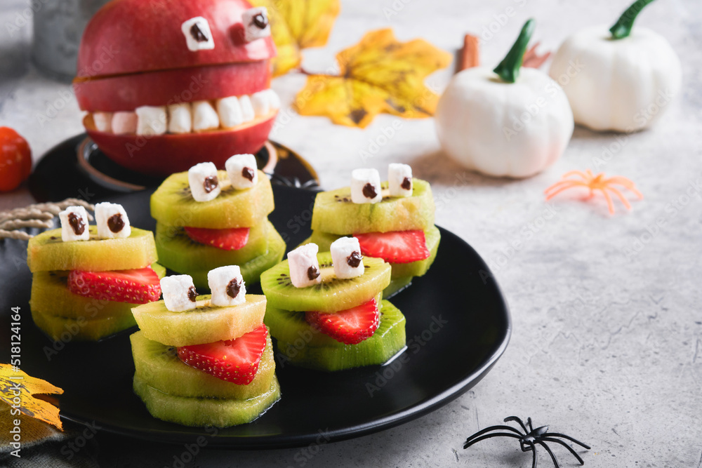Spooky green kiwi monsters for Halloween. Healthy Fruit Halloween Treats. Halloween party kiwi, strawberry, apple and marshmallow monster on grey stone or concrete table background. Selective focus.