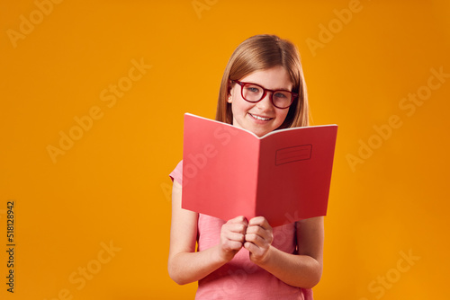 Studio Shot Of Young Girl Studying School Exercise Book Wearing Glasses Against Yellow Background