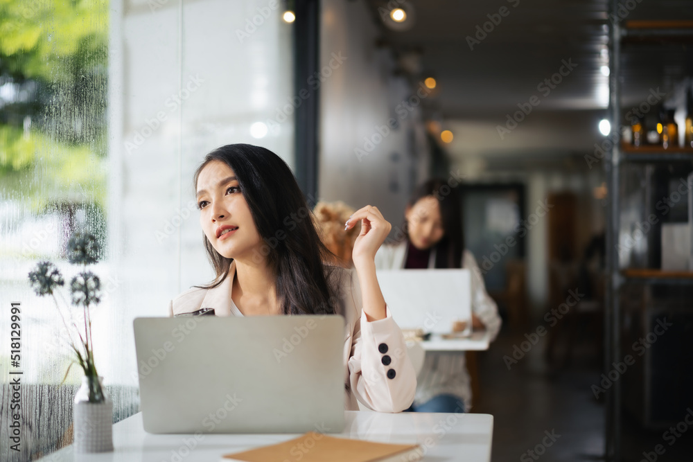 Asian woman using laptop in restaurant and looking outside dreaming about something