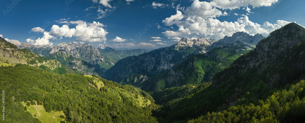 A panoramic, aerial view of the monumental Albanian Alps. Hiking trails, dark blue sky with clouds, steep rocks, green valley, remnants of snow, summer. Theth National Park, Albanian Alps, Albania.