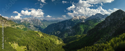 A panoramic, aerial view of the monumental Albanian Alps. Hiking trails, dark blue sky with clouds, steep rocks, green valley, remnants of snow, summer. Theth National Park, Albanian Alps, Albania.