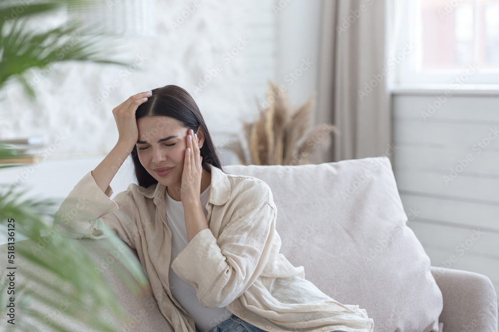 Young upset woman alone at home, depressed woman sitting on sofa in living room, holding hand to head, grimacing in pain and depression Young upset woman alone at home, depressed woman sitting on sofa in living room, holding hand to head, grimacing in pain and depression