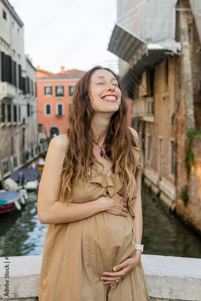 Happy pregnant woman touching belly in blurred Venice.