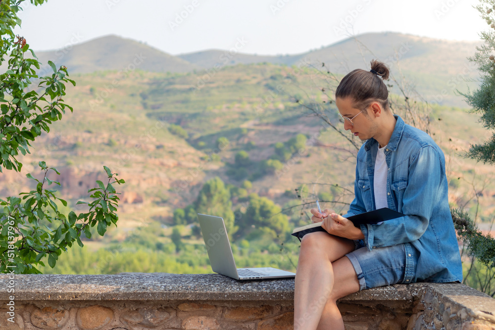 Young man working as a freelancer on his computer and drawing in his notebook surrounded by a natural landscape