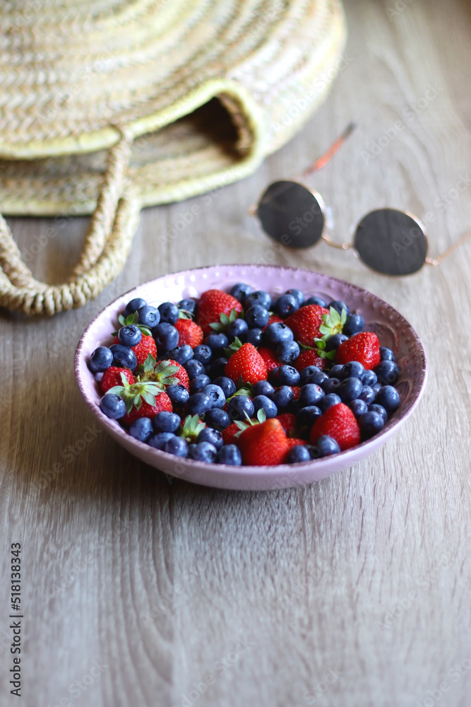 Bowl of strawberries and blueberries, sunglasses and wicker bag on the wooden table. Selective focus. Bowl of strawberries and blueberries, sunglasses and wicker bag on the wooden table. Selective focus.