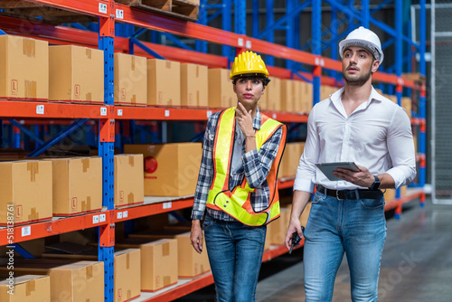 Latino executive and a young Latino employee wearing safety jackets and helmets talk to each other, checking inventory in a warehouse. © Supavadee