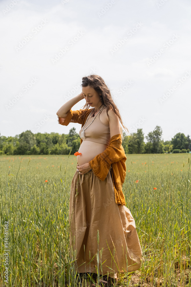 Naklejka premium Pregnant woman holding pappy flower while standing in field.