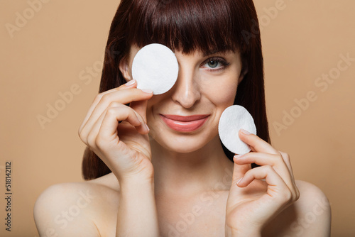 Joyful young female covering eye with cotton pad posing and having fun caring for face skin standing on beige background, smiling looking at camera. Studio shot. Facial skincare concept. photo