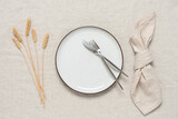 Autumn table setting. A white plate, cutlery, a beige linen napkin and ripe ears of wheat on a beige linen tablecloth. Top view, flat lay.
