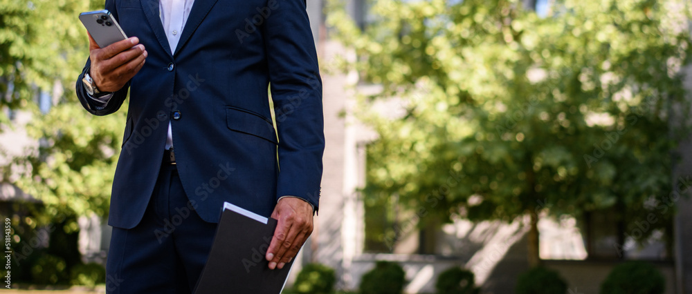 Faceless shot of african american businessman using smartphone and holding paper folder outdoors, banner crop Faceless shot of african american businessman using smartphone and holding paper folder outdoors, banner crop