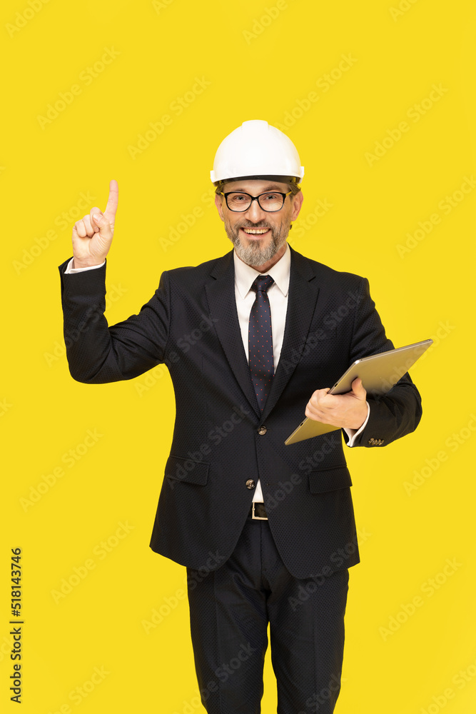 Handsome middle aged businessman in black suit pointing up holding digital tablet smiling looking at camera isolated on yellow background. Businessman in suit construction hard hat and eye glasses. Handsome middle aged businessman in black suit pointing up holding digital tablet smiling looking at camera isolated on yellow background. Businessman in suit construction hard hat and eye glasses.