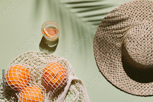 Summer flat lay with straw hat, glass of water and orange fruit in eco shopping bag. Green background with palm leaf shadow, sun and sunlight. Vacation, holiday, summer creative minimal concept.