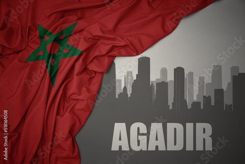 abstract silhouette of the city with text Agadir near waving colorful national flag of morocco on a gray background.