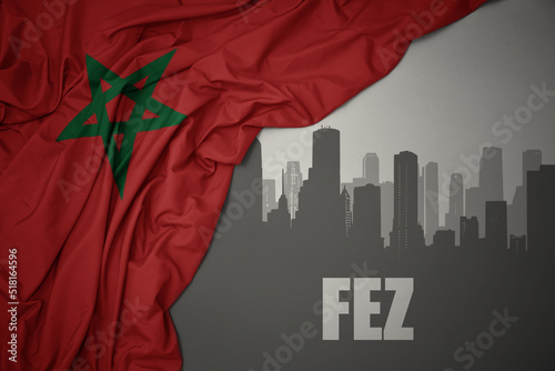 abstract silhouette of the city with text Fez near waving colorful national flag of morocco on a gray background.