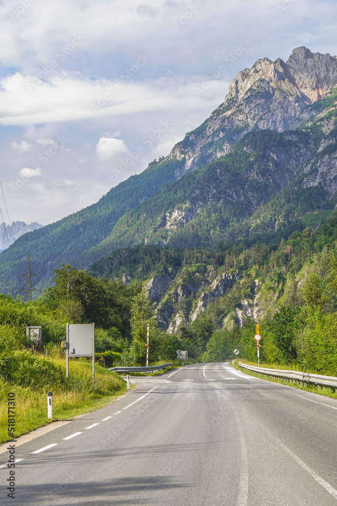 Beautiful road to the Alps in Austria leading to wooded slopes against a blue sky with clouds.
