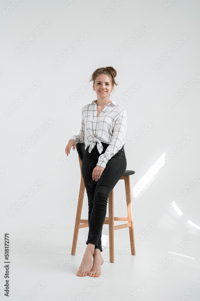 Fototapeta premium A sexy slender girl with hair in a bun is stylishly dressed in a shirt and pants, isolated on grey background. A woman sits on a high chair in a photo studio