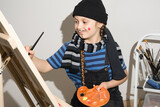 Child artist concentrates painting a picture on an easel by paints, a brush and an keep orange palette in her hands. On the girl's face, a neat, thin black French mustache is drawn. View is straight.
