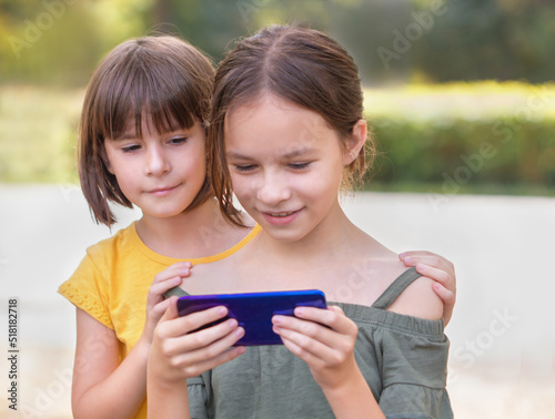 Two happy girls looking at cellphone. Pretty children surfing internet at the park