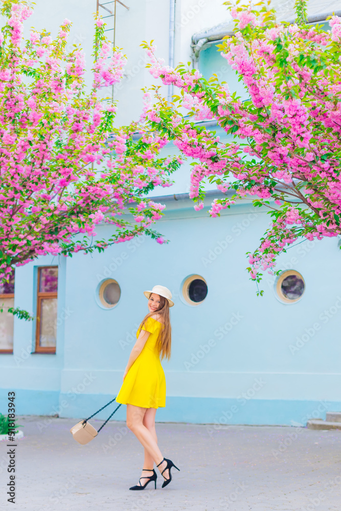 Φωτογραφία Girl in yellow dress and hat holds a handbag next to the building and branches o