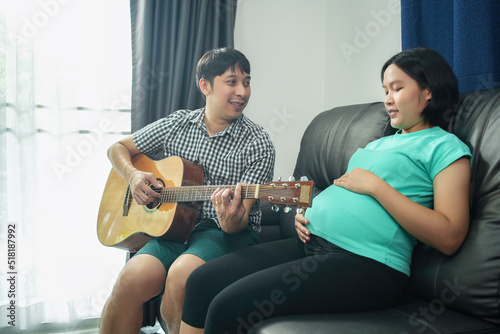 Asian husband and pregnant wife spend time together in the bedroom and husband play guitar sing-song for an unborn baby at home.