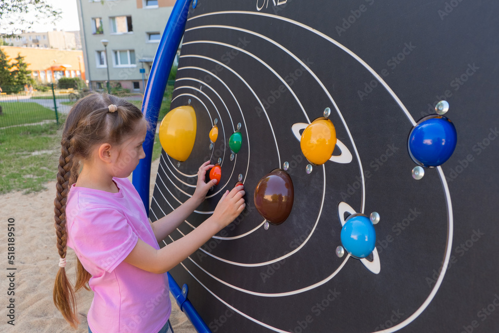 Fototapeta premium A little girl of 7-8 years old with long hair, dressed in a pink T-shirt and shorts, studies the planets of the star system located on the board in an amusement park.