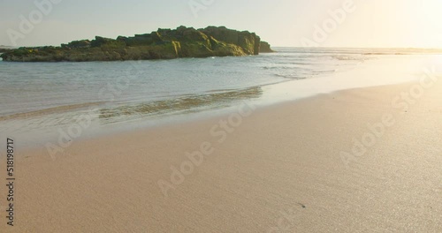 Rocky shore with green seaweed on stones, low tide, golden sandy beach. Calm ocean waves in the background. Sunset on the island Fuerteventura. Magic soft light, slow motion. Static shot. No people.