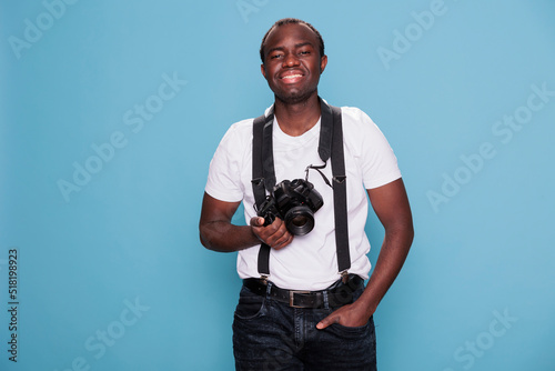 Confident photographer having professional camera smiling heartily while standing on blue background. Handsome looking young man wearing fashion clothes while having photo device.