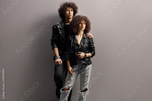 Young man and woman with leather jackets and curly hair leaning on a gray wall