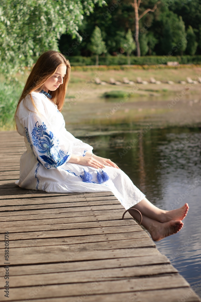 a girl in an embroidered Ukrainian shirt sits on the pier, the reflection of clouds in the water of the lake. On the shore of the sky. vyshyvanka day. freedom. patriot