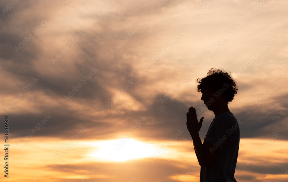 Silhouette of a christian man praying with the sunset in the background. Person thanking God.