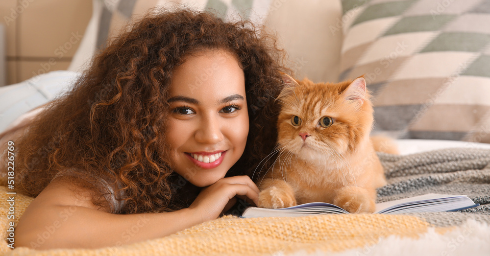 Young African-American woman with cute cat reading book in bedroom