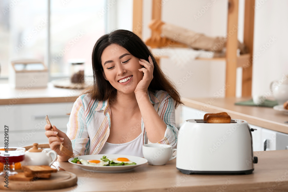 Beautiful young Asian woman drinking coffee and eating tasty toasts with fried eggs in kitchen Beautiful young Asian woman drinking coffee and eating tasty toasts with fried eggs in kitchen