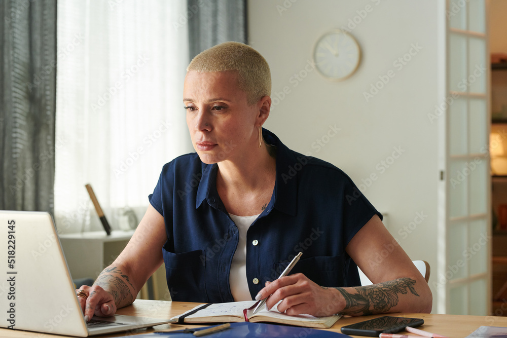 Serious woman using her laptop and making notes in notepad while sitting at table and working online from home