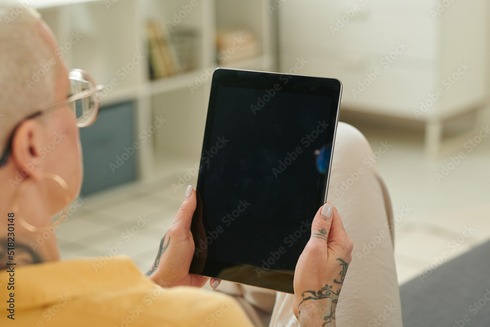 Rear view of woman in eyeglasses holding digital tablet in front of her and talking online or watching video while resting at home