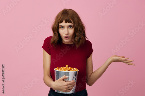 Excited shocked cute redhead lady in red t-shirt with popcorn ready for movie evening posing isolated on over pink studio background. Copy space Banner. Fashion Cinema concept. Entertainment offer