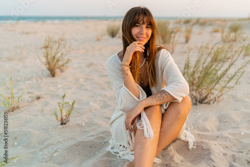 Smiling pretty woman in bohemian summer outfit with stylish neacklase  sitting on san , enjouing  warm summer sunset.  Looking on camera. photo